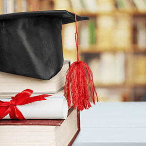 Mortar board with tassel sits on top of stacked books next to a diploma