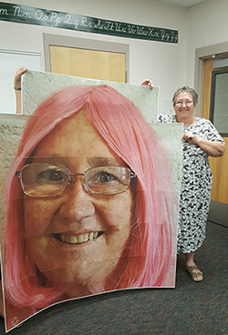 Staff member poses with a large poster of Principal Debbie Mulder in a pink wig