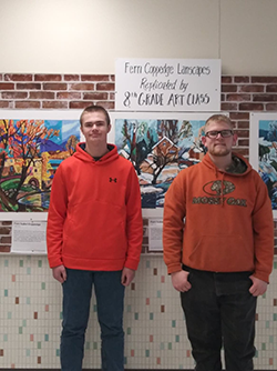 Oliver Johnson and Owen Holmberg pose together in front of a wall of art and a sign reading Fern Coppedge Landscapes Replicated by 8th Grade Art Class